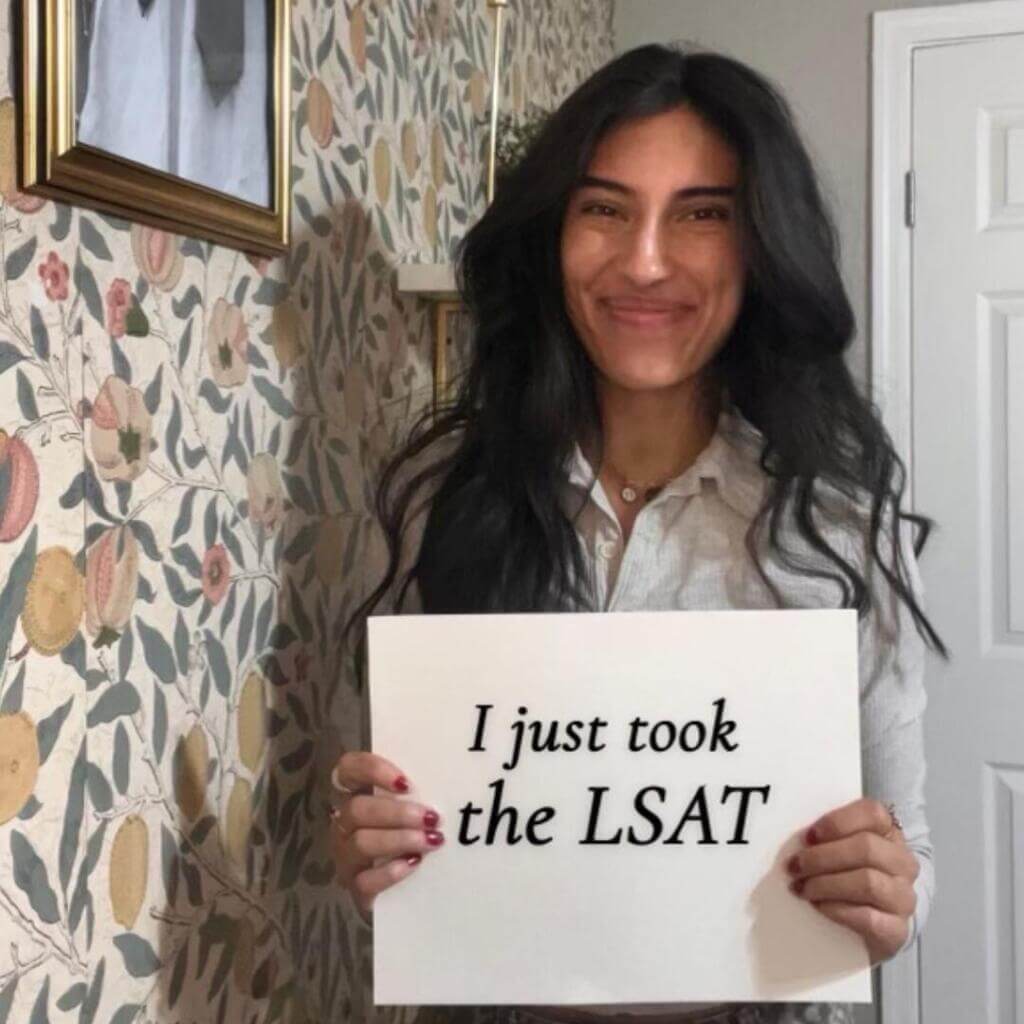 A woman stands indoors against a floral wallpaper holding a sign that reads "I just took the LSAT" with a proud smile on her face.