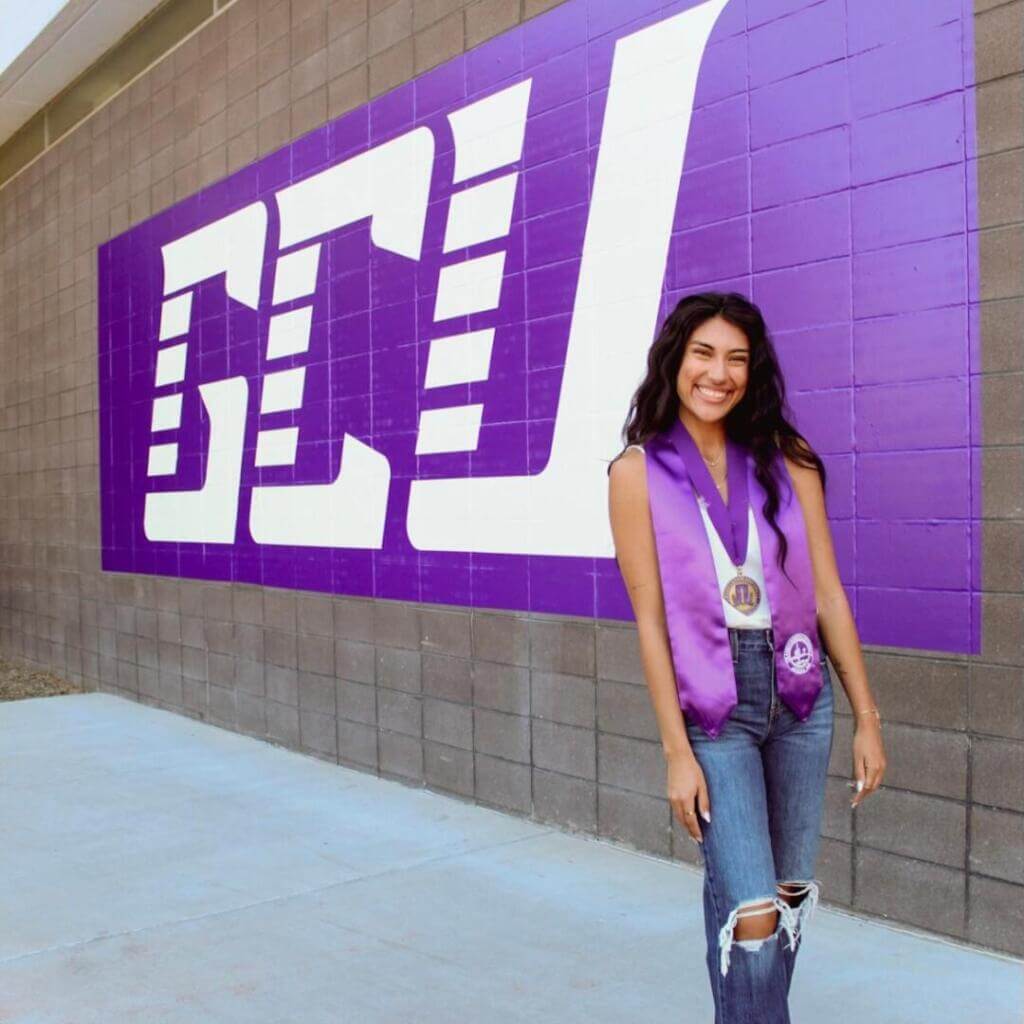 A young woman wearing graduation regalia stands in front of a purple "GCU" wall mural, smiling confidently. She wears a medal and a lavender stole.