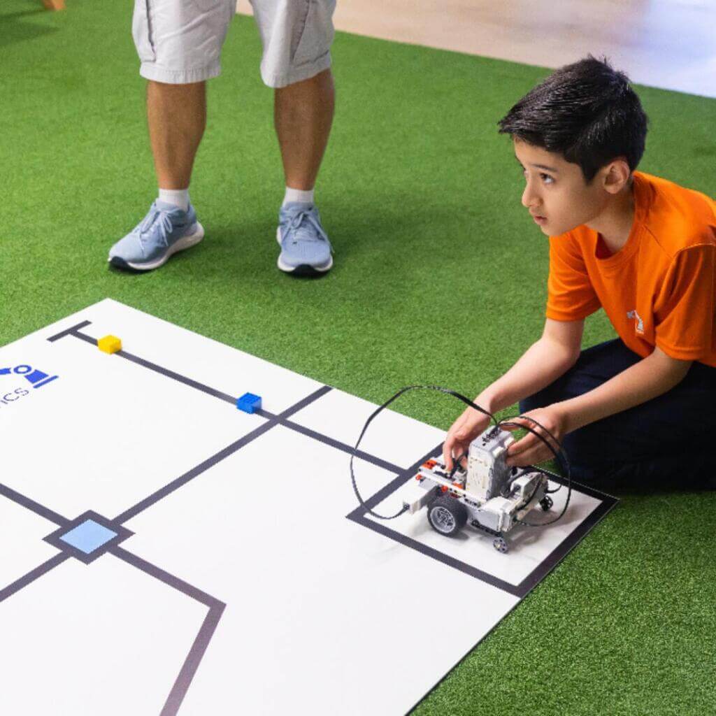 A young boy in an orange shirt kneels on artificial grass while operating a wheeled robot on a printed course mat. An adult’s legs are visible nearby, suggesting supervision or instruction.