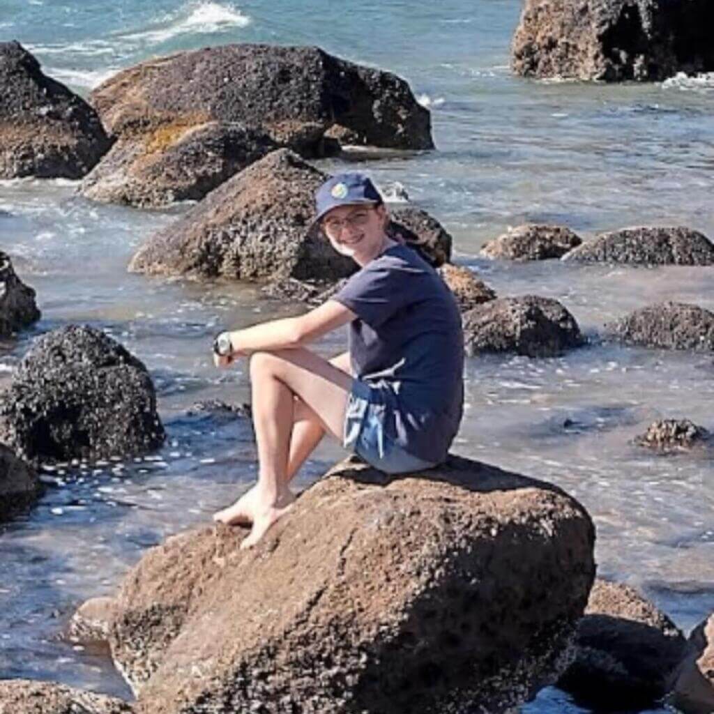 A barefoot child in a navy outfit and cap sits on a large rock surrounded by ocean water and other rocks, smiling while enjoying a sunny day at the shore.