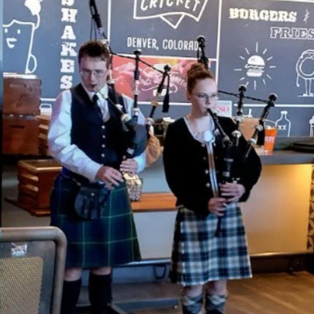 Two girls and one boy dressed in traditional Highland attire smile for a photo indoors. The girls wear colorful kilts and black jackets, while the boy wears a black jacket, blue kilt, and a beret.