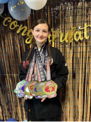 Person wearing a martial arts uniform holding multiple championship medals with a congratulatory backdrop and balloons.