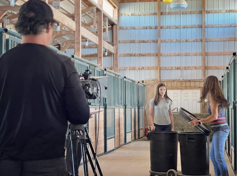 Indoor barn setting with two people near large bins and a person filming with a camera on a tripod.