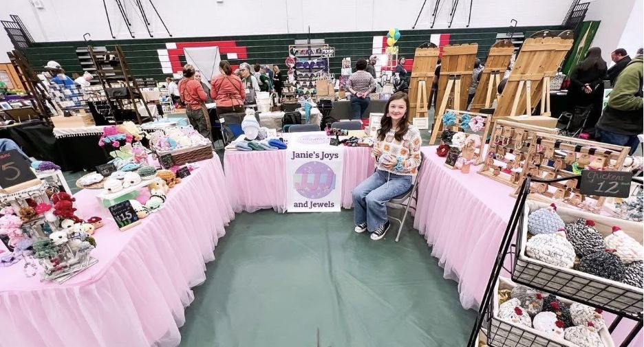 Craft fair booth with pink table covers displaying handmade items, labeled “Janie’s Joys and Jewels,” with a person seated at the center.