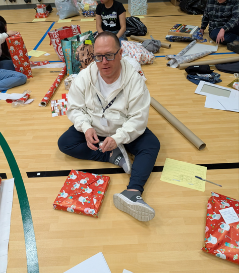 A person sits on a gymnasium floor surrounded by rolls of wrapping paper, tape, and wrapped gifts, working on wrapping a present with red holiday-themed paper.