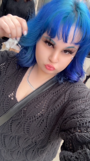 A person with bright blue hair takes a selfie while wearing a black sweater and necklace.