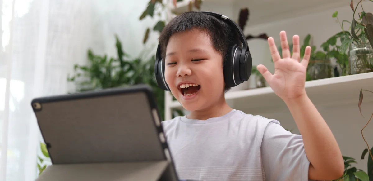 Elementary student waiving at his online class wearing a black pair of headphones