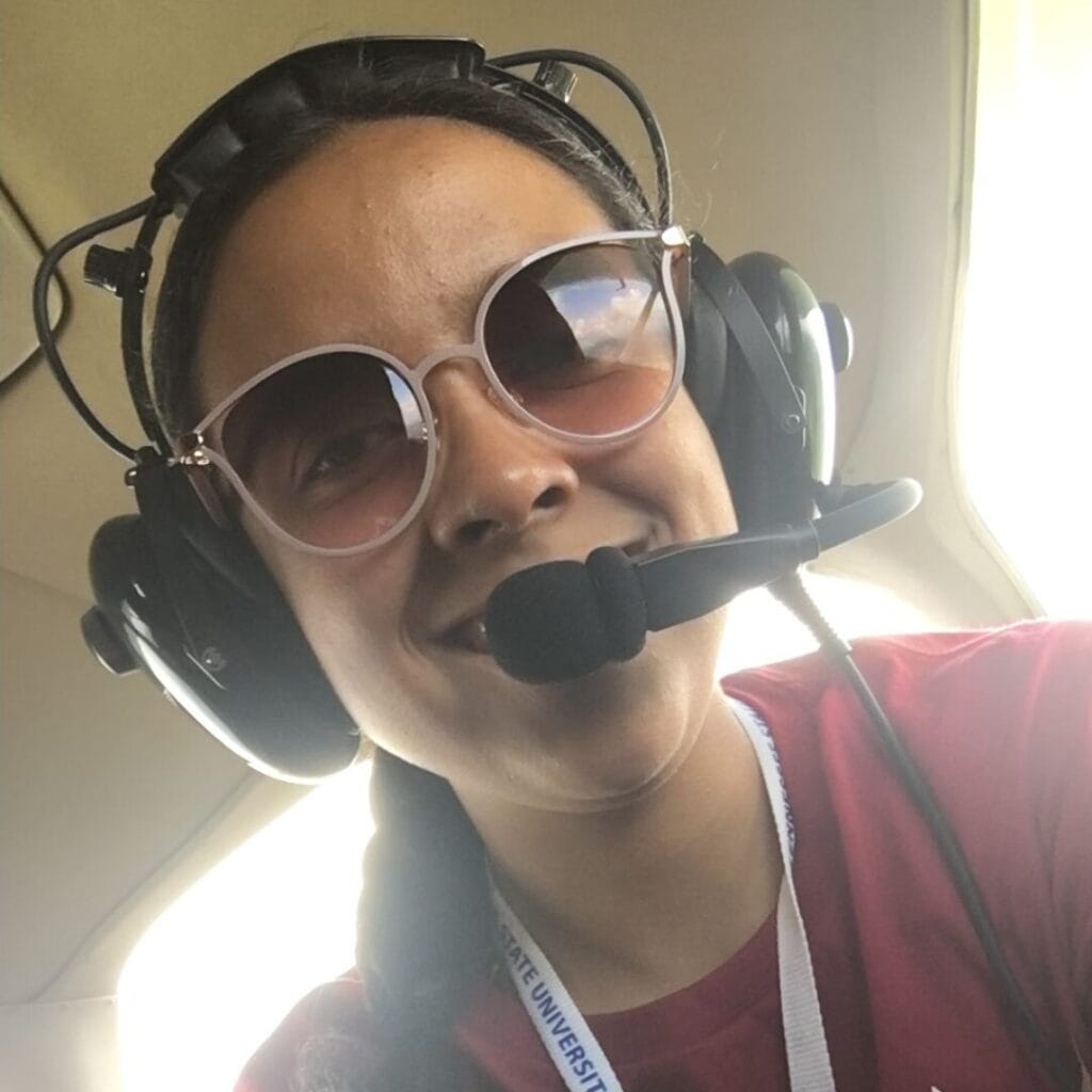 Person wearing aviator sunglasses and a headset with a microphone, smiling inside an aircraft.