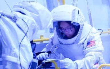 Two astronauts in spacesuits are working on equipment inside a training facility