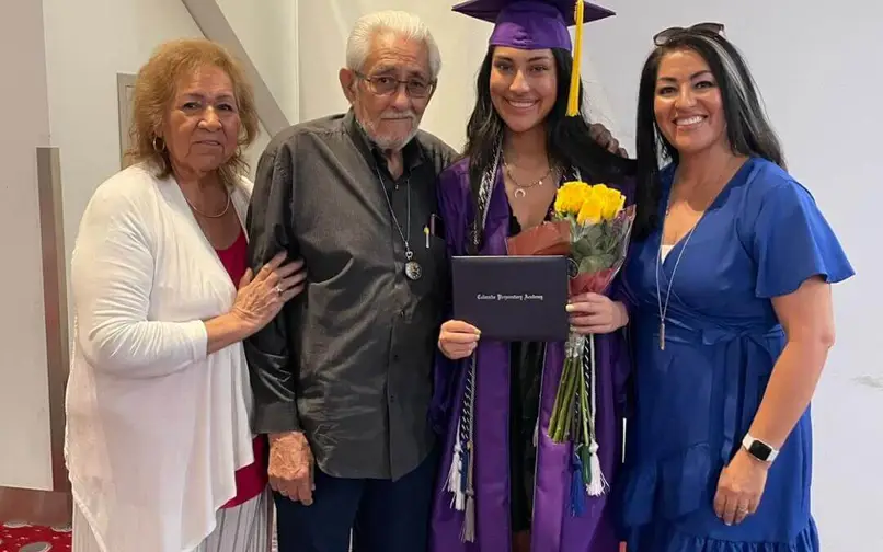 A graduate in a purple cap and gown holds a diploma and yellow flowers while standing with three smiling family members, including two women and an elderly man.
