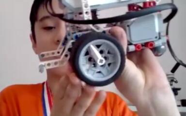 A boy wearing an orange shirt and a medal holds up a LEGO-style robot, showing its wheel and wiring up close, proudly displaying his project.