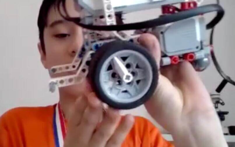 A boy wearing an orange shirt and a medal holds up a LEGO-style robot, showing its wheel and wiring up close, proudly displaying his project.