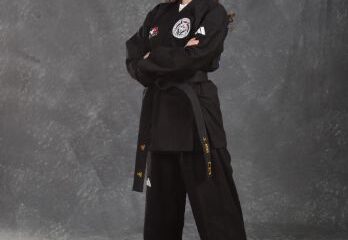 Person standing barefoot in a black martial arts uniform with a black belt, arms crossed, against a gray studio background.