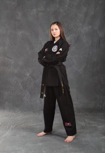 Person standing barefoot in a black martial arts uniform with a black belt, arms crossed, against a gray studio background.
