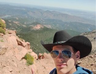Person wearing a cowboy hat and blue hoodie giving a thumbs-up on a rocky mountain summit with expansive forested valleys in the background.