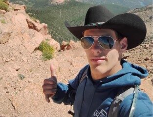 Person wearing a cowboy hat and blue hoodie giving a thumbs-up on a rocky mountain summit with expansive forested valleys in the background.