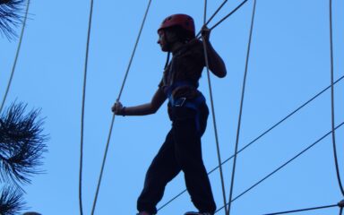 A participant wearing a helmet and safety harness walks across a suspended rope bridge at an outdoor ropes course.