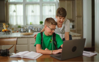 A student sits at a kitchen table working on a laptop with open textbooks, while an adult stands nearby offering support in a home learning environment.