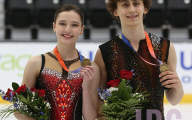 Two figure skaters wearing gold medals hold bouquets and pose on the ice after a competition.