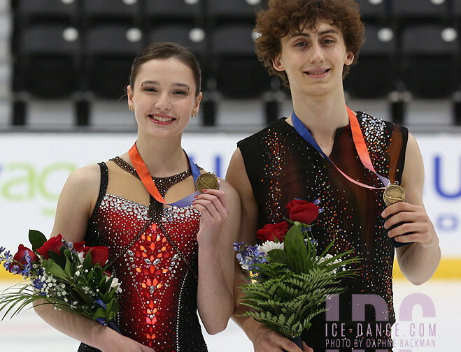 Two figure skaters wearing gold medals hold bouquets and pose on the ice after a competition.